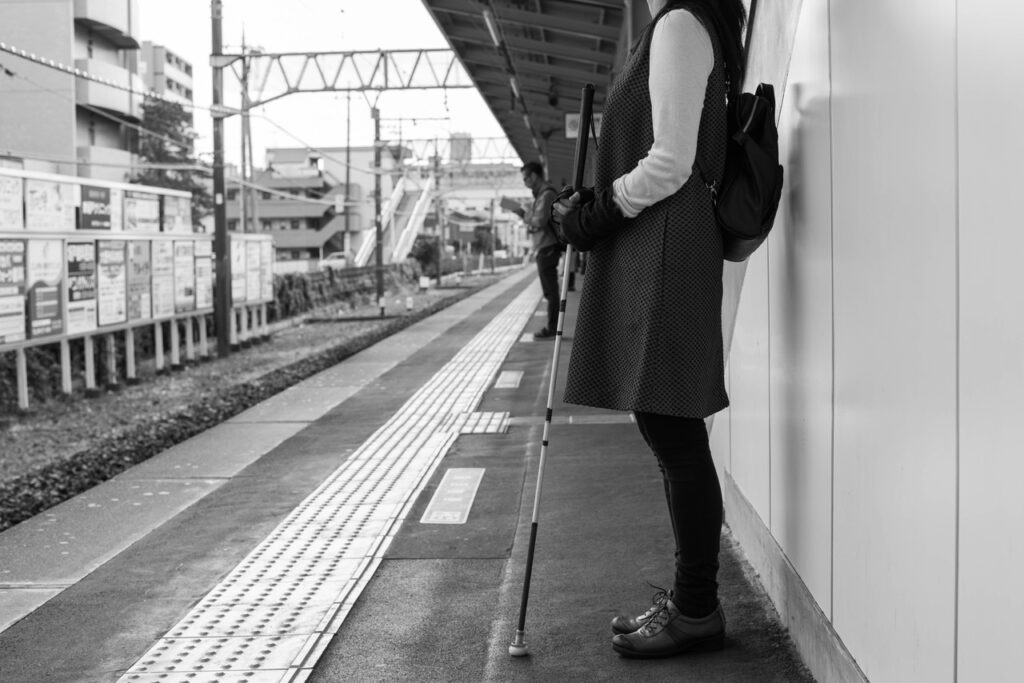 白杖を持った人が駅のホームで電車の到着を待っている写真