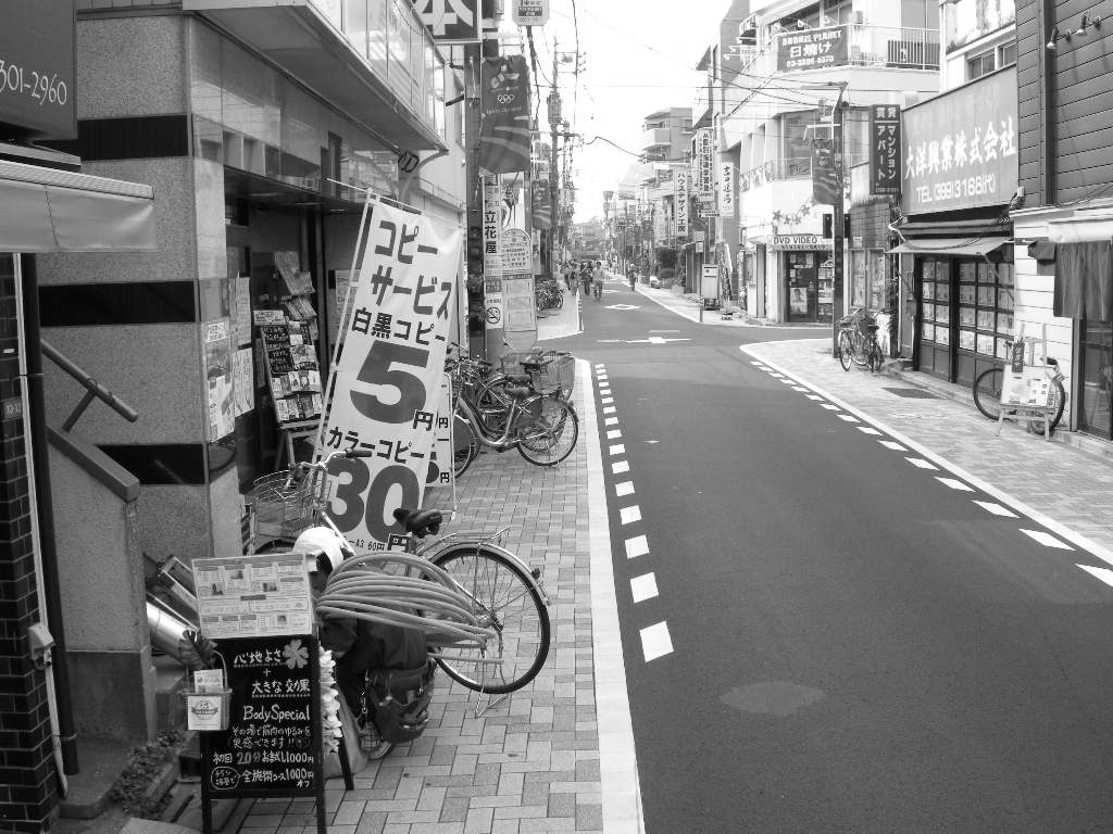 商店街の店の前に看板や自転車が乱雑に置かれているモノクロ写真。