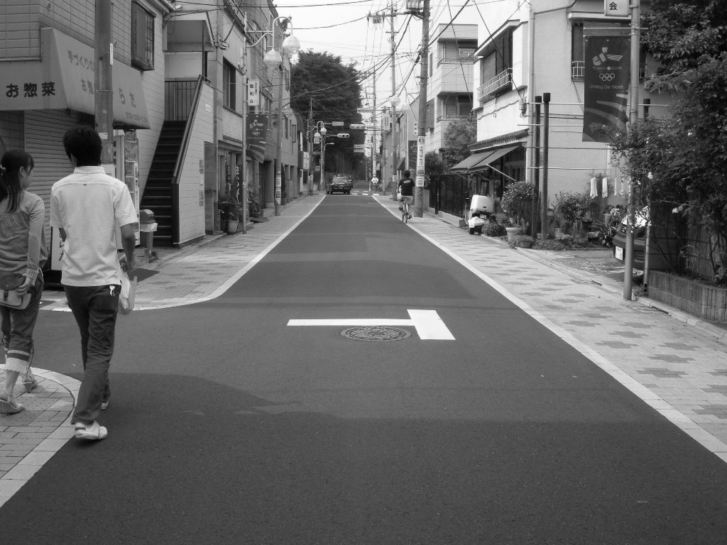 道の両側に店が点在し男女二人連れの歩行者、道の先に走っている自転車と自動車が写っている写真