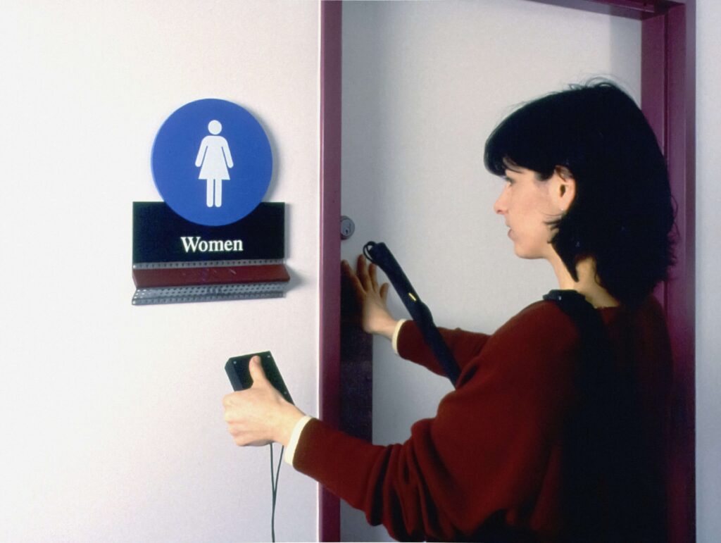 視覚障害のある女性が白杖を右脇に挟み左手でトイレ表示にポケットサイズの電子機器を向け、ドアを押してトイレに入ろうとしている写真