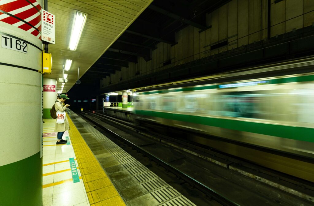 夜の駅のプラットホームの写真。点字ブロックの前で電車の到着を待っている人が一人いる。向かい側の線路を電車が通過している。