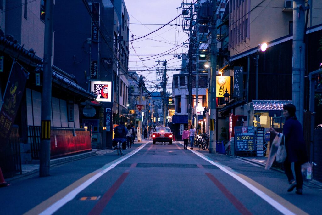 夕方の街中の写真。道路の両脇に路側帯があり、車道を一台のタクシーが走り歩道を数名の歩行者が歩いたり自転車に乗っている人がいる。