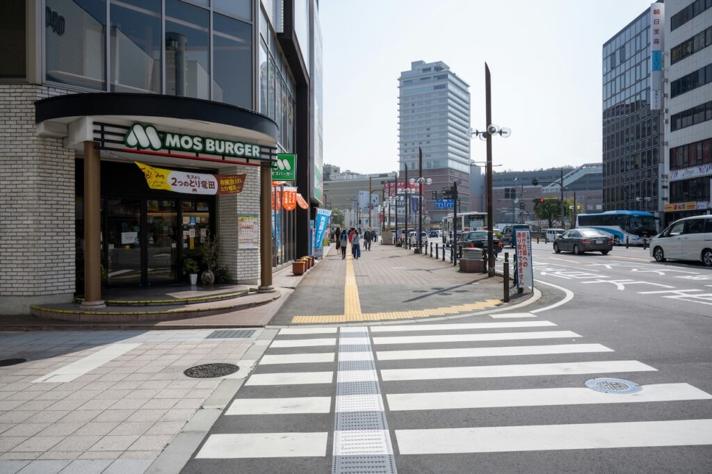 ファーストフード店の前の交差点の写真。店の前の歩道の角が大きく丸くなっていて、そこから横断歩道が始まっている。その丸みに沿うように点字ブロックが敷設されている。横断歩道上にはエスコートゾーンが敷設されている。