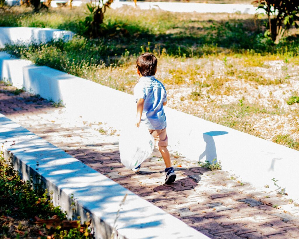 4歳くらいの男の子が大きなビニール袋を左手に持ってレンガ敷の歩道を歩いている後ろ姿の写真。歩道の両脇には白いコンクリートの縁石があり左が側は下、右側は上に段差になっている)、その脇には雑草が生えている。