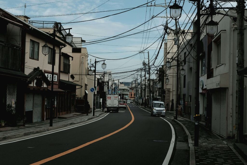 街中の二車線道路。前方に向かって走行している車が2台、対向車線には路駐している車が一台写っている。道路の両脇には低層の店、住宅が密集していて電柱が多く立っている。