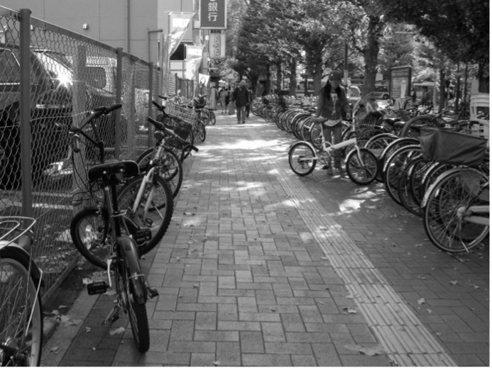 車道側に駐輪スペースがあり、民地側にも自転車が多数停められている歩道の写真。歩道の真ん中に点字ブロックが敷設されている。点字ブロックのすぐ脇に一台の自転車の後輪がはみ出ている。