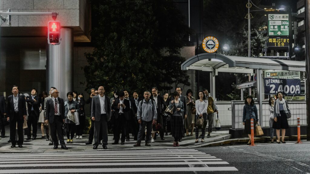夜、仕事帰りの人々が横断歩道の前で信号待ちをしている写真。