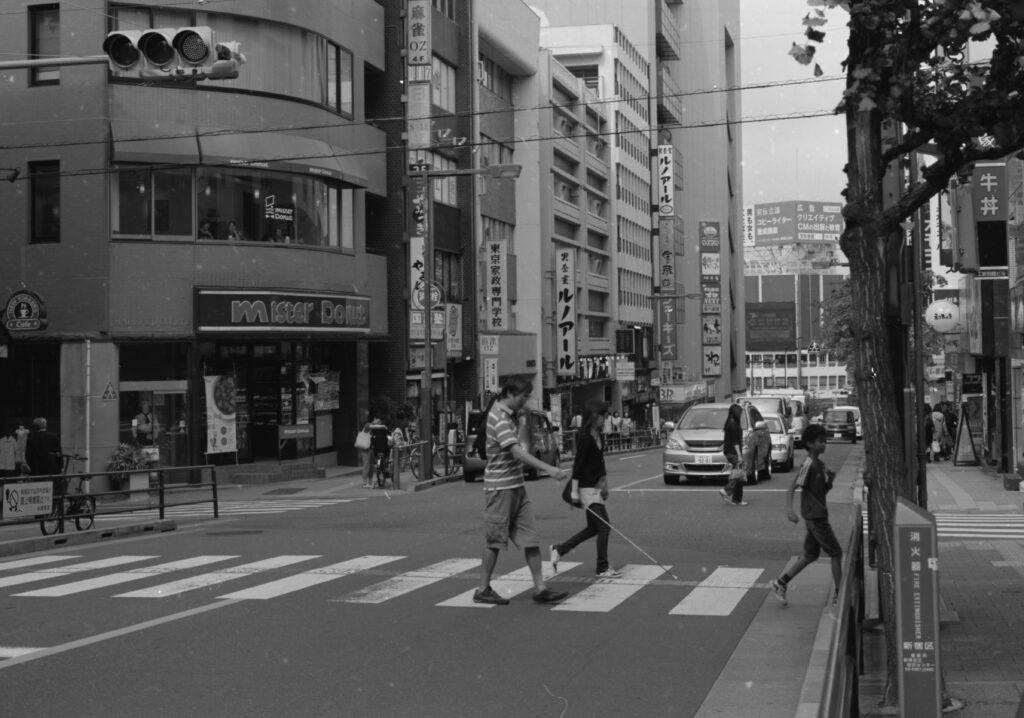 店が立ち並ぶ街中の横断歩道を白杖を持った男性が歩いている写真。近くに女性や子どもが同じ方向に横断している。車道には車が数台停止して信号が変わるのを待っている。