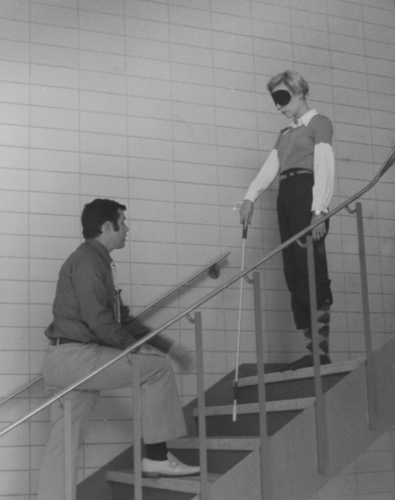 アイマスクをつけた金髪の女性が白杖を使って階段を降りようとしている写真。階段の3段下に男性の訓練士段を跨いで指導している。